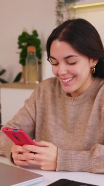 Evde cep telefonu kullanan mutlu genç bir kızın cep telefonuyla cep telefonu uygulamasında sosyal medya içeriğinin keyfini çıkardığı dikey HD videosu. Teknoloji yaşam tarzı konsepti