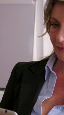Profesyonel orta yaşlı iş kadınının dijital tablette gezinen, şirket ofisinde çalışırken dikkatle konsantre olan, doğal pencere ışığıyla aydınlatılan dikey HD videosu