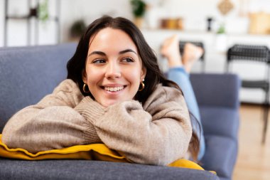 Huzurlu genç bir kadın oturma odasında rahat bir kanepede uzanıp huzur ve mutluluğun tadını çıkarıyor.