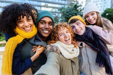 Çeşitli mutlu genç arkadaşlar kış tatilinde selfie çektirirken eğleniyorlar. Çok ırklı genç insanlar dışarıda kendi portrelerinde kameraya gülümsüyor. Gençlik toplumu.