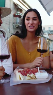 Restoran masasının önünde kırmızı şarapla aperatif yiyen üç genç kadının dikey HD videosu. Kadın arkadaşlığı ve tatil yaşam tarzı konsepti.