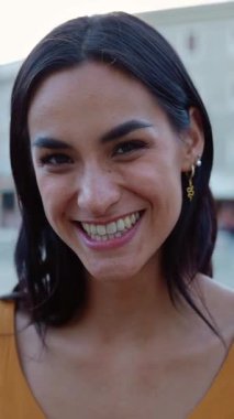 Dikey HD video. Şehir caddesinde kameraya gülümseyen beyaz güzel bir kadının yakın plan portresi. Mutluluk ve pozitif insanlar kavramı.