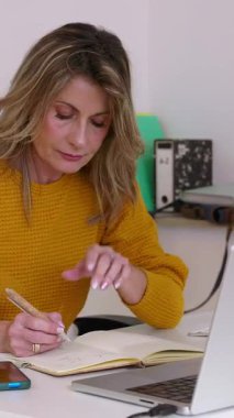 Profesyonel iş kadınının özenle ev ofisinde çalıştığı, notlar aldığı ve dizüstü bilgisayarda yazarak üretkenliğini ve stratejik düşüncelerini gösterdiği dikey HD videosu