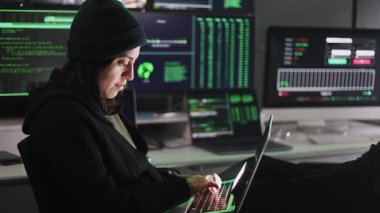 Arka planda birden fazla monitör olan karanlık odada siber saldırı için dizüstü bilgisayarda virüs kodları yazan profesyonel bir kadın hacker.