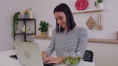 Mutfakta taze salatanın tadını çıkarırken dizüstü bilgisayarda çalışan mutlu genç kadın. Teknoloji ve ev hayatı kavramı