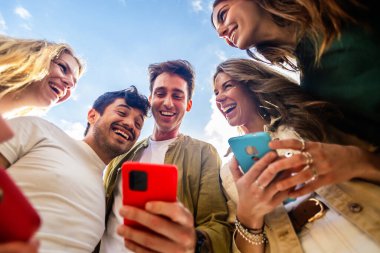 Bir grup genç cep telefonu kullanıyor, mavi gökyüzüne karşı sosyal medyanın tadını çıkarıyor. Sosyal medya ve teknoloji yaşam tarzı kavramı