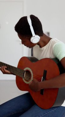 Gülümseyen Afrikalı genç bir kadının evdeki dizüstü bilgisayarda çevrimiçi ders izlerken akustik gitar çaldığı dikey HD video. E-öğrenme, eğitim ve çevrimiçi sınıf kavramı