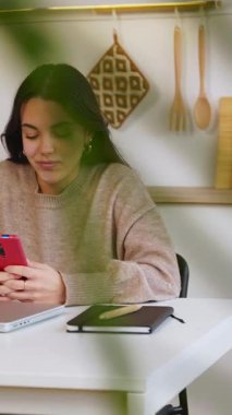 Evdeki mutfak masasında oturan, sosyal medyanın içeriğini izleyen veya arkadaşlarıyla online sohbet eden mutlu genç bir kadının dikey HD videosu