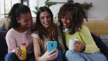 Üç neşeli genç kadın akıllı telefonlarıyla selfie çekerken evde rahat bir kanepede oturup meyve suyu içerken komik surat ifadeleri takınıyorlar.