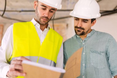 Güvenlik kaskı ve yüksek görüş açısına sahip yelek giyen iki mühendis planları gözden geçirip inşaat alanında proje detaylarını tartışıyorlar. Mühendislik ve mimari kavramı