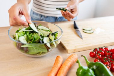 Genç bir kadın modern bir mutfakta sağlıklı salata hazırlıyor, salatalık ve marul kasesine taze salatalık ekliyor. Sağlıklı yaşam tarzı ve gıda konsepti