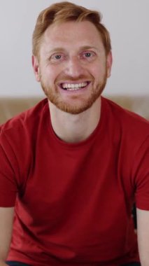 Dikey video. Yeni dairede otururken kameraya bakan mutlu genç adamın portresi. Mutlu insanlar, yeni hayat ve başlangıç konsepti