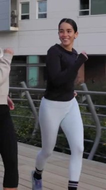 Üç genç kız arkadaşın birlikte koştuğu, kentsel parkta ağaçlar ve köprüyle koşarken gülümseme enerjisini paylaştığı dikey HD videosu fitness arkadaşlığını temsil ediyor.