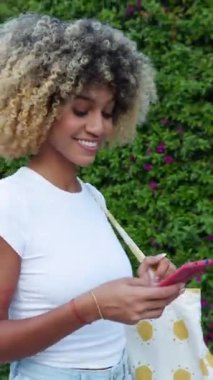 Dikey HD video. Genç Afro-Amerikan bir kadının şehir caddesinde cep telefonu kullanışının yan görüntüsü. Z Gen kızı yazın dışarıda akıllı telefon uygulamasından mesaj yolluyor. Teknoloji yaşam tarzı konsepti