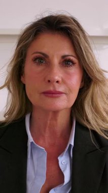 Ofiste kameraya gülümserken masada oturan orta yaşlı iş kadınının dikey HD video portresi. Güzel, olgun bir menajerin yakın plan portresi.