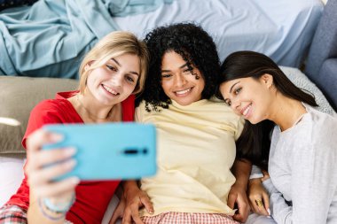 Üç neşeli çoklu etnik kız bir pijama partisi sırasında yatakta uzanırken akıllı telefonuyla selfie çekiyor. Kadın arkadaşlığı ve gençlik kavramı