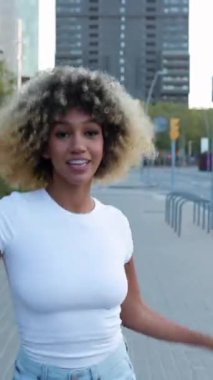 City Street 'te dans eden mutlu Latin Amerikalı kadının dikey HD videosu. Binlerce yıllık etnik kızın kameraya doğru giderken telefonda dans müziğinin tadını çıkarmasını ön planda görmek. Mutluluk kavramı