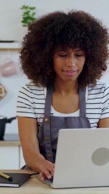 Mutfakta yemek pişirirken dizüstü bilgisayarda çalışan mutlu genç kadının dikey HD videosu. Seyahat ve iş konsepti