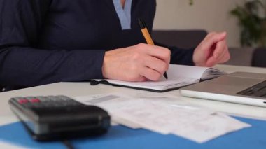 Bilgisayarı ve hesap makinesiyle bir ofis masasında otururken not defterine not alan tanınmayacak kadar olgun bir iş kadını.