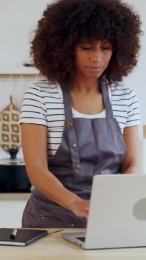 Dikey HD video. Arkadan, genç, çok ırklı bir kadının tencereyi karıştırması, evden çalışırken modern mutfağında sağlıklı bir yemek pişirmesi. Çoklu görev ve tele çalışma kavramı