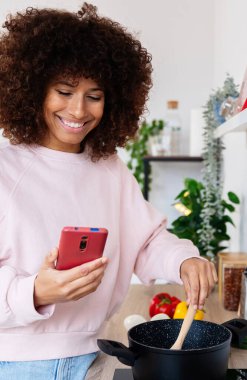 Gülümseyen, güzel, çok kültürlü bir kadın akıllı telefona bakıyor, tencereyi karıştırıyor, modern mutfağında sağlıklı bir yemek pişiriyor, çorba hazırlıyor.