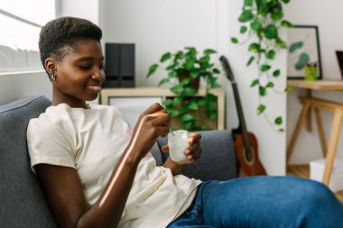 Huzurlu genç kadın rahat bir ev ortamında rahat kanepesinde sağlıklı bir yoğurt atıştırıyor.