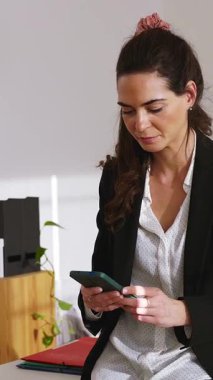 Profesyonel iş kadınının masada otururken akıllı telefonuyla mesajlaştığı, çağdaş kurumsal çalışma alanında doğal ışıklandırmayla mutluluk saçan dikey HD videosu.