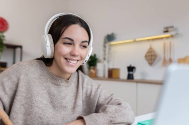 Kulaklık takan mutlu öğrenci çevrimiçi öğrenim için dizüstü bilgisayar kullanıyor, mutfakta kahvaltıdan keyif alıyor. Eğitim ve e-öğrenim kavramı