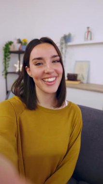 Evdeki kanepede dinlenirken cep telefonuyla video görüşmesi yapan mutlu genç bir kadının dikey HD videosu. İletişim konsepti.