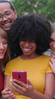 Şehir caddesinde cep telefonuyla gülen bir grup gencin dikey HD videosu. Milenyum kuşağı arkadaşları sosyal medya uygulamasında içeriği izlerken eğleniyorlar.