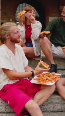 Şehrin caddesinde margherita pizza yiyen genç insanların dikey HD videosu. Milenyum turist arkadaşları İtalya 'da yaz tatilinin tadını çıkarırken kasaba meydanında fast food yiyorlar.