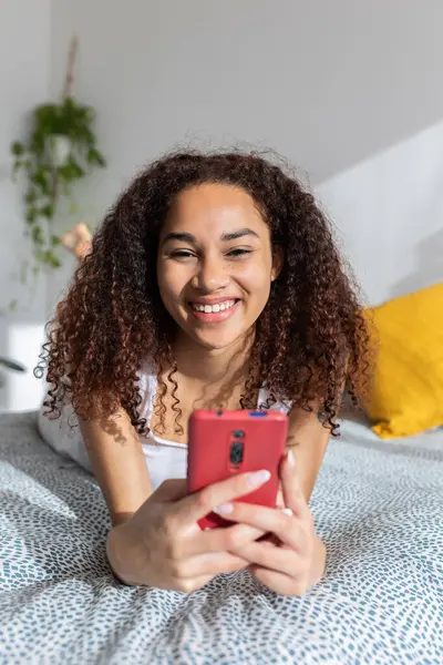 Genç Latin bir kadın rahat bir yatakta uzanıyor, evde akıllı telefonunu mutlu bir şekilde kullanıyor, rahat bir sabahın tadını çıkarırken sosyal medyada geziniyor.