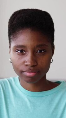 Evdeki oturma odasında kameraya bakan genç, ciddi siyahi bir kadın. Gençlik, güven ve Afrikalı insanlar konsepti. Dikey HD görüntüler