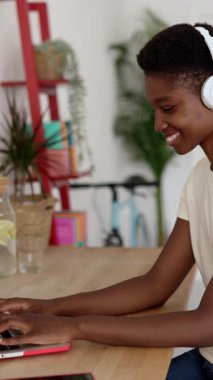 Genç ve mutlu bir Afrikalı kadının dizüstü bilgisayar kullandığı dikey HD video. Girişimci kadın stüdyo işyerinde çalışırken kulaklıkla müzik dinliyor..