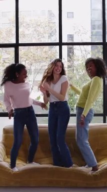 Evdeki kanepede dans eden, partiden keyif alan, birlikte kutlama yapan üç mutlu genç kızın dikey HD videosu.