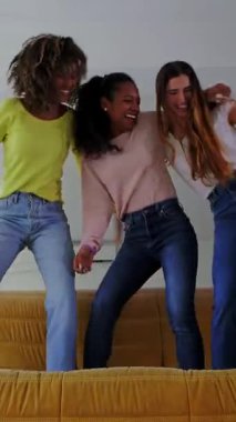 Üç yetişkin kadının evdeki kanepede dans edişinin dikey HD videosu. Birleşik çok ırklı kadınlar kanepeye atlıyor. Arkadaşlık ve mutluluk kavramı. 