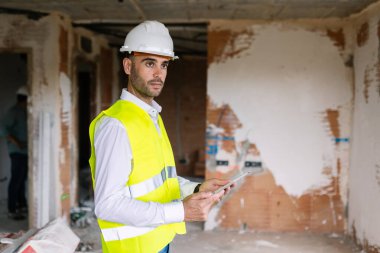 Güvenlik yeleği ve kask giyen profesyonel mühendis tablet kullanıyor, inşaat alanının çalışmalarını denetliyor. Mühendislik ve mimari kavramı