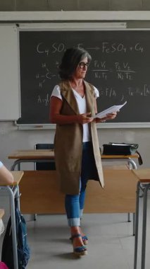 Yetişkin bir bayan öğretmenin sınıfta lise öğrencilerine sınav dağıtırken çekilmiş dikey HD videosu. Eğitim yaşam tarzı kavramı.