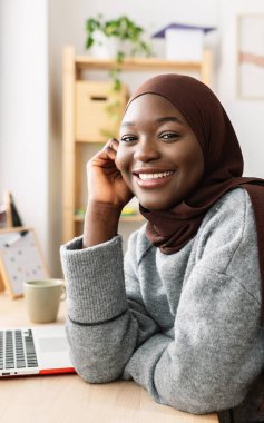 Masada oturan kameraya gülümseyen Afrikalı genç bir kadının dikey portresi. Müslüman başörtüsü takmış mutlu siyah kız öğrenci evde dinleniyor. Gençlik ve etnik toplum kavramı.