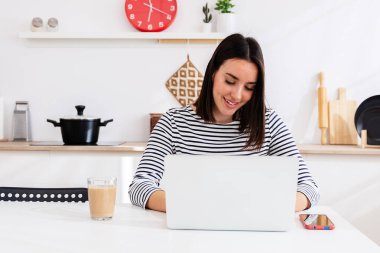Genç mutlu kadın sabahları modern mutfakta bilgisayarla kahve ve akıllı telefonla çalışıyor.