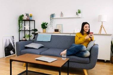 Kafkasyalı mutlu bir kadın koltukta cep telefonu kullanarak rahatlıyor. Milenyum kadını elinde cep telefonu cihazıyla kanepede dinleniyor, sosyal medya ağlarından içeriği izliyor.