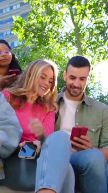 Şehirde cep telefonu kullanan genç bir grubun dikey HD videosu. Gülümseyen çok ırklı arkadaşlar dışarıda oturmuş cep telefonu uygulamasında sosyal medya içeriğini izliyorlar.