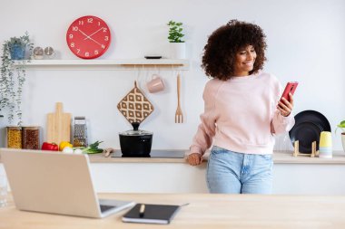 Genç ve mutlu bir kadın cep telefonu kullanırken bilgisayarla geziyor ve modern mutfakta yemek pişiriyor. İş ve teknoloji kavramı