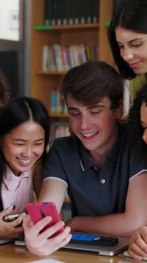 Lise kütüphanesinde ders çalışmaya ara verirken sosyal medya içeriğini izlerken eğlenen bir grup öğrencinin dikey HD videosu. Teknoloji ve eğitim yaşam tarzı konsepti.