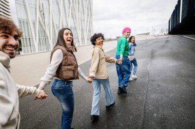 Çeşitli genç arkadaşlar el ele tutuşurken birlikte mutlu bir şekilde koşuyorlar. Gençlik toplumu, arkadaşlık ve birlik kavramı