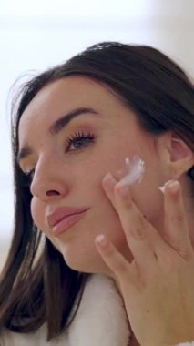 Güzel bir kadının yüzüne nemlendirici krem sürerken banyoda cilt bakımının keyfini çıkarırken çekilmiş dikey HD videosu.