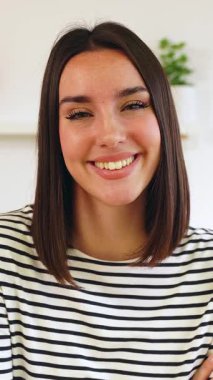 Evde kameraya gülümseyen mutlu genç bir kadının dikey HD video portresi.