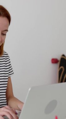 Evde dizüstü bilgisayarda çalışan güzel mutlu genç kadının dikey HD videosu. Milenyum dişisi oturma odasındaki masada bilgisayar başında oturuyor..