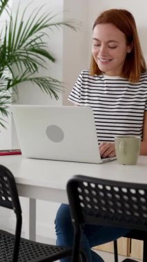 Evde dizüstü bilgisayarda çalışan gülümseyen genç yetişkin bir kadının dikey HD videosu. Oturma odasındaki bilgisayarla çalışan beyaz bir kadın. Uzak çalışma ve eğitim kavramı.