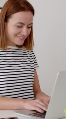 Oturma odasındaki masada oturan ve dizüstü bilgisayarla çalışan neşeli genç kadının dikey HD videosu. Milenyum kız öğrencisi evden online olarak çalışıyor. Teknoloji yaşam tarzı ve eğitim kavramı.